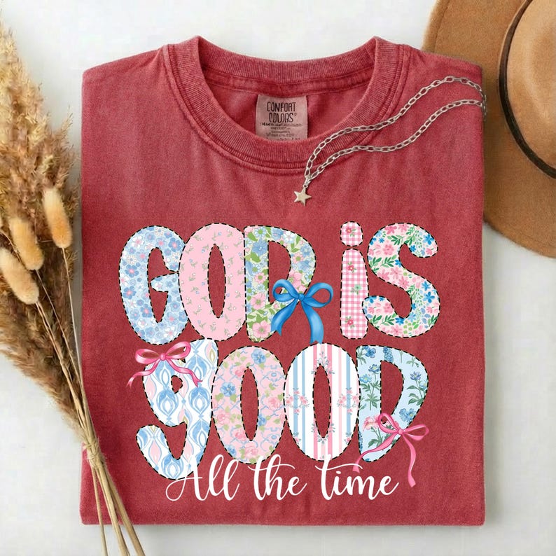 Puede incluir: Una camiseta roja con la frase "GOD IS GOOD" en un dise&ntilde;o de patchwork con estampados florales y a rayas. La frase "All the time" est&aacute; debajo. Un collar plateado con un colgante de estrella est&aacute; sobre la camiseta.