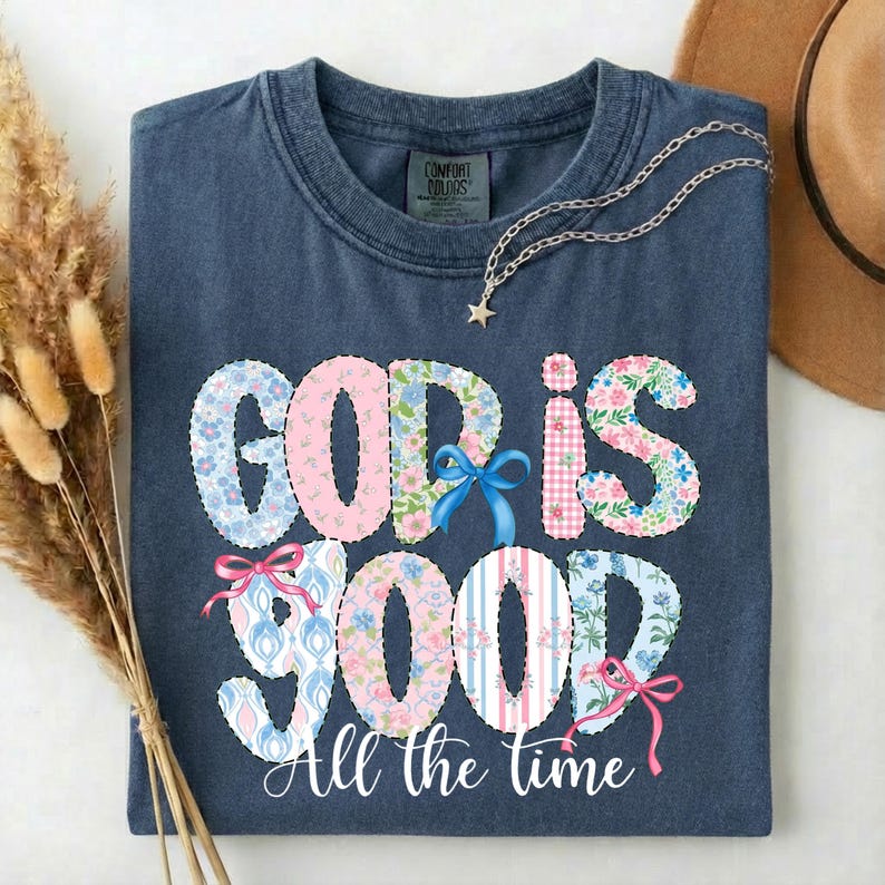 Puede incluir: Una camiseta azul marino con las palabras "GOD IS GOOD" en un dise&ntilde;o de patchwork con estampados florales y a rayas. La frase "All the time" est&aacute; escrita debajo en una fuente cursiva. Un collar plateado con un colgante de estrella descansa sobre la camiseta.