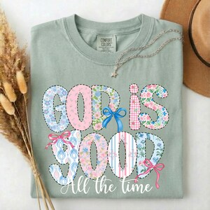 Puede incluir: Camiseta verde salvia con las palabras "GOD IS GOOD" en un dise&ntilde;o de patchwork con estampados florales y a rayas. La frase "All the time" est&aacute; escrita debajo. Se ve un collar plateado y un sombrero marr&oacute;n.