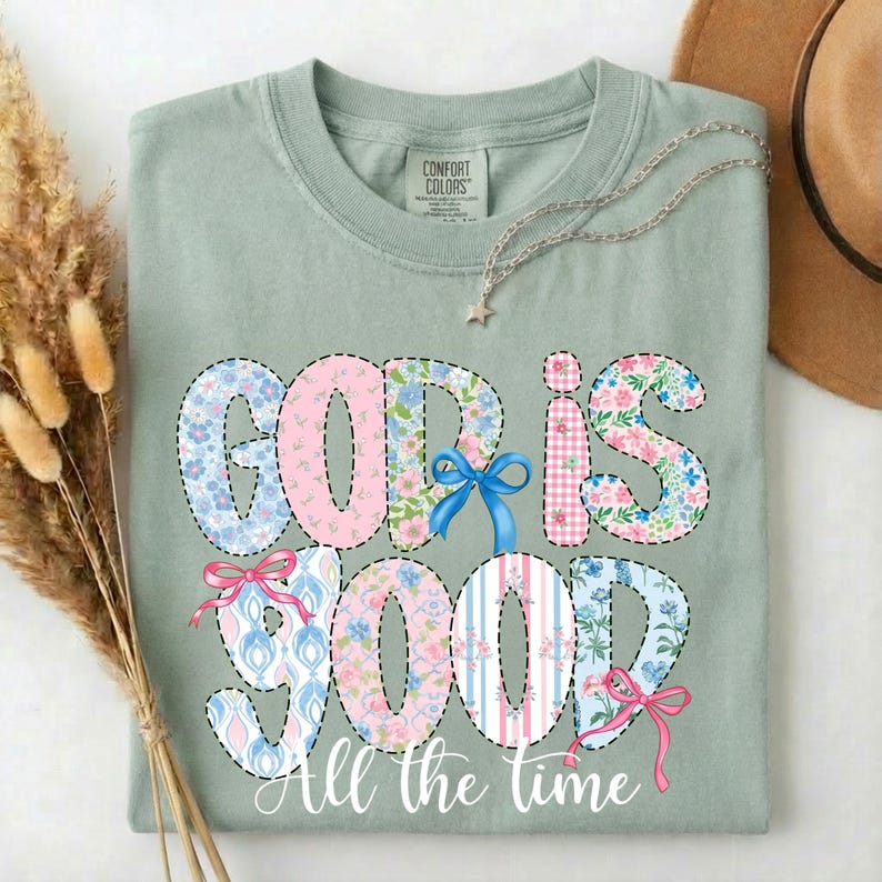 Puede incluir: Camiseta verde salvia con las palabras "GOD IS GOOD" en un dise&ntilde;o de patchwork con estampados florales y a rayas. La frase "All the time" est&aacute; escrita debajo. Se ve un collar plateado y un sombrero marr&oacute;n.