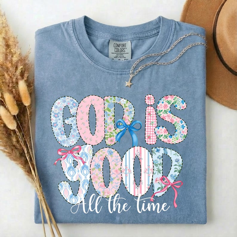 Puede incluir: Una camiseta azul claro con las palabras "GOD IS GOOD" en un dise&ntilde;o de patchwork. Las letras presentan telas florales, a rayas y estampadas, con lazos. La frase "All the time" est&aacute; escrita debajo.