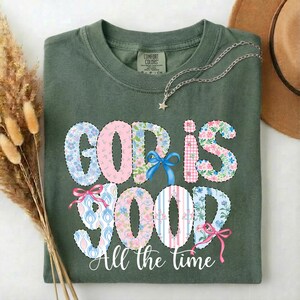 Puede incluir: Camiseta verde oliva con la frase "GOD IS GOOD" en un dise&ntilde;o floral y estampado. La frase "All the time" est&aacute; escrita debajo en una fuente cursiva. Un collar plateado con un colgante de estrella est&aacute; sobre la camiseta.