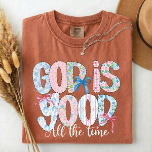 Puede incluir: Una camiseta de color &oacute;xido con las palabras "GOD IS GOOD" en un dise&ntilde;o de patchwork. Las letras est&aacute;n decoradas con estampados florales, rayas y lazos. La frase "All the time" est&aacute; escrita debajo.
