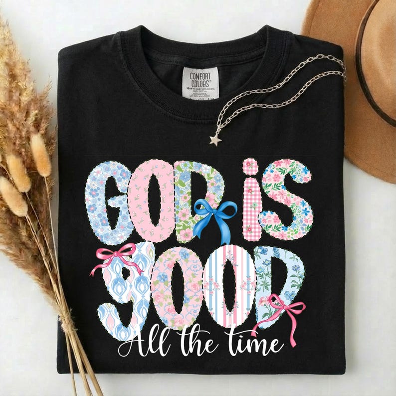 Puede incluir: Camiseta negra con la frase "GOD IS GOOD" en un dise&ntilde;o floral y estampado. La frase "All the time" est&aacute; impresa debajo. Se ve un collar plateado con un colgante de estrella y un sombrero marr&oacute;n.