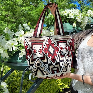 Peut inclure: Un sac fourre-tout à motifs blancs, rouges et noirs, avec une fermeture à cordon. Le sac est doté d'une large bandoulière à rayures rouges et noires. Le sac est présenté sur un fond de feuillage vert et de fleurs blanches.