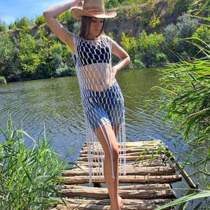 May include: A woman wearing a straw cowboy hat, a white net top, and denim shorts stands on a wooden dock near a river. The white net top has a honeycomb design with long fringe. The background includes green trees and water.