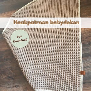 Puede incluir: Una manta de bebé de crochet beige con un borde blanco. La manta está hecha con un patrón de punto de waffle. El texto "Haakpatroon babydeken" está escrito en la parte superior de la manta. El texto "PDF Download" está escrito en un círculo verde en la esquina inferior izquierda de la manta.