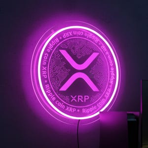 Könnte beinhalten: Ein leuchtendes, pinkfarbenes XRP Ripple Coin Neon-Schild. Das runde Schild zeigt das XRP-Symbol in der Mitte und die Worte "Ripple coin XRP" am Rand. Es ist an einer dunklen Wand über einem Schreibtisch angebracht.