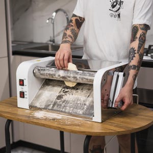 May include: A white pasta roller machine with a red digital display and a person rolling out dough. The machine has a lever on the side and a roller in the centre.