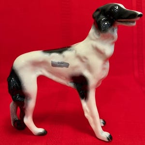 May include: A black and white porcelain figurine of a dog, standing on all four legs. The dog is a Greyhound breed and has a black spot on its back. The figurine is labeled "Duke 2031".