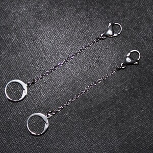 May include: Two silver-coloured chain extenders with lobster claw clasps and circular ring attachments. The chains are thin and delicate, designed for jewellery or accessories. The background is a dark, textured surface.