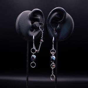 May include: A pair of silver-toned earrings displayed on black ear-shaped stands. Each earring features a silver chain, circular metal rings, and a dark, multi-coloured bead with blue and brown swirls. One earring includes a connecting chain.