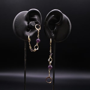 May include: A pair of gold-toned earrings featuring a chain design with purple spherical beads and small clear accents. One earring connects two hoops with a short chain, while the other features a longer chain drop ending in a single hoop.