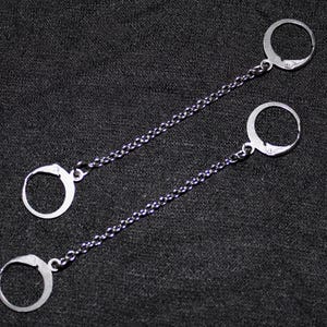 May include: A pair of silver earrings with a chain design. Each earring has a circular hoop attached to a delicate chain, with another hoop at the opposite end. The earrings are set against a dark background.