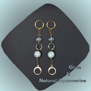 Pode incluir: Um par de brincos de argola dourados com contas penduradas de água-marinha e vidro. Os brincos são exibidos em uma superfície cinza escuro. O texto "Glass & Natural Aquamarine" é visível.