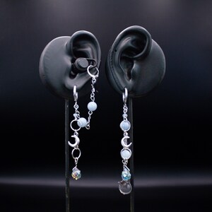 May include: A pair of silver-toned chain earrings featuring light blue spherical beads, small silver crescent moon charms, and faceted crystal teardrop pendants. One earring includes a secondary chain connecting to a small hoop for a double piercing.