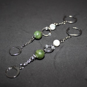 May include: Two silver finger rings connected by a chain to a ring. Each chain has a green and white bead. The background is a dark grey.