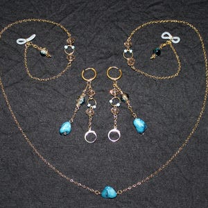 Puede incluir: Un conjunto de joyas de color dorado que incluye un collar, pendientes y cadenas para gafas. El collar y las cadenas para gafas tienen un diseño de cadena con un colgante de piedra azul. Los pendientes tienen un diseño de aro con cuentas colgantes y una piedra azul.