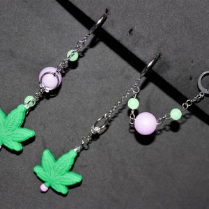 May include: Dangle earrings with green cannabis leaf charms and pink details. The earrings have silver-tone chains, lavender and green beads, and lever-back closures. The earrings are displayed on a dark grey surface.
