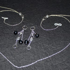 May include: A silver jewellery set comprising two necklaces and a pair of earrings. The necklaces have fine chains with black and clear beads. The earrings have a similar design, with black beads and dangling clear beads. The jewellery is displayed on a dark background.
