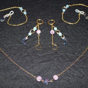 May include: A gold-coloured jewellery set featuring a necklace, two pairs of earrings, and two eyeglass chains. The necklace and chains have pink and blue beads. The earrings have pink beads, gold hoops, and crescent moon charms.