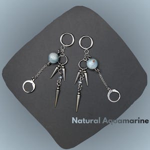 Può includere: Un paio di orecchini argentati a tema acquamarina naturale. Ogni orecchino presenta un piccolo cerchio, una perla azzurra e una catena con un piccolo ciondolo circolare. Sotto la perla ci sono tre punte argentate. Il testo "Natural Aquamarine" è visibile.