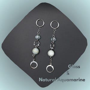 May include: Dangling earrings with silver hoops and chains. Each earring has a large, round, off-white bead, smaller clear beads, and a small silver hoop at the bottom. The text "Glass & Natural Aquamarine" is visible.