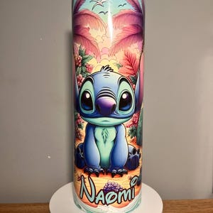 Op de afbeelding: Een blauw en turquoise beker met het personage Stitch uit Lilo & Stitch. Het ontwerp bevat een tropische strandsc&egrave;ne met palmbomen, bloemen en de naam "Naomi". De beker is cilindrisch en lijkt van roestvrij staal te zijn.