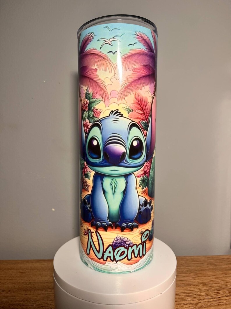 Op de afbeelding: Een blauw en turquoise beker met het personage Stitch uit Lilo & Stitch. Het ontwerp bevat een tropische strandsc&egrave;ne met palmbomen, bloemen en de naam "Naomi". De beker is cilindrisch en lijkt van roestvrij staal te zijn.