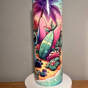 Op de afbeelding: Een roestvrijstalen beker met een kleurrijke strandsc&egrave;ne, inclusief een paarse palmboom, surfplanken en het personage Stitch. Het ontwerp bevat het woord "mi" en is geplaatst tegen een blauwe lucht en oceaan.