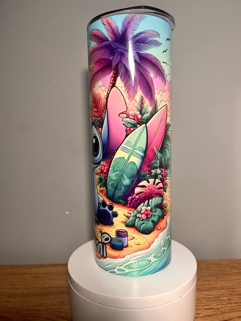 Op de afbeelding: Een roestvrijstalen beker met een kleurrijke strandsc&egrave;ne, inclusief een paarse palmboom, surfplanken en het personage Stitch. Het ontwerp bevat het woord "mi" en is geplaatst tegen een blauwe lucht en oceaan.