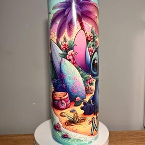 Op de afbeelding: Een roestvrijstalen beker met een levendige illustratie van een stripfiguur op een tropisch strand. Het ontwerp omvat een surfplank, palmboom en bloemen, met de naam van het personage op het zand. De beker is cilindrisch en heeft een blauw deksel.
