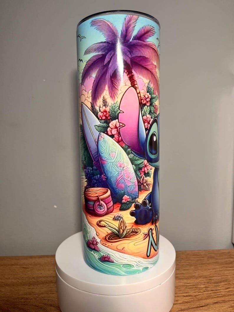 Op de afbeelding: Een roestvrijstalen beker met een levendige illustratie van een stripfiguur op een tropisch strand. Het ontwerp omvat een surfplank, palmboom en bloemen, met de naam van het personage op het zand. De beker is cilindrisch en heeft een blauw deksel.