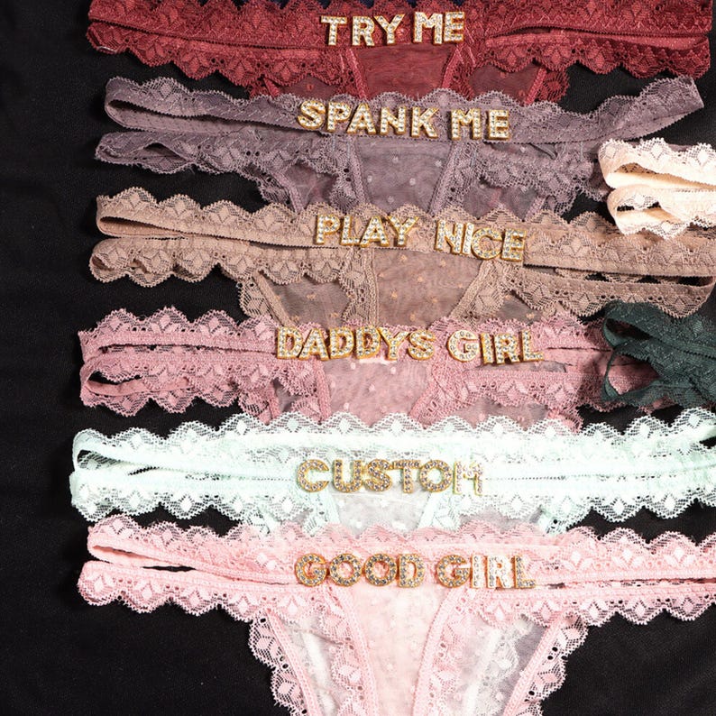 K&ouml;nnte beinhalten: Eine Kollektion aus sechs Spitzen-Tangas in verschiedenen Farben wie Burgunderrot, Mauve, Beige, Altrosa, Mintgr&uuml;n und Hellrosa. Jedes Paar ist mit goldenen Strasssteinen verziert, die die Schriftz&uuml;ge TRY ME, SPANK ME, PLAY NICE, DADDYS GIRL, CUSTOM und GOOD GIRL bilden.