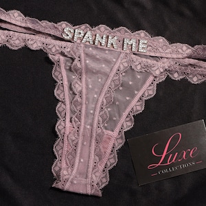 May include: Mauve thong underwear with lace trim and a sheer dotted panel. The waistband features the text "SPANK ME" in silver rhinestones. A black card with the text "Luxe Collections" is also present.