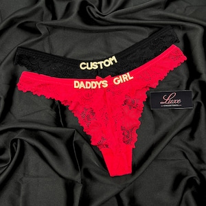 Pode incluir: Um par de tangas de renda, uma preta e uma vermelha, com letras douradas. A pe&ccedil;a preta exibe a palavra CUSTOM, enquanto a pe&ccedil;a vermelha exibe o texto DADDYS GIRL. Uma pequena etiqueta preta com o texto branco Luxe COLLECTIONS &eacute; vis&iacute;vel.
