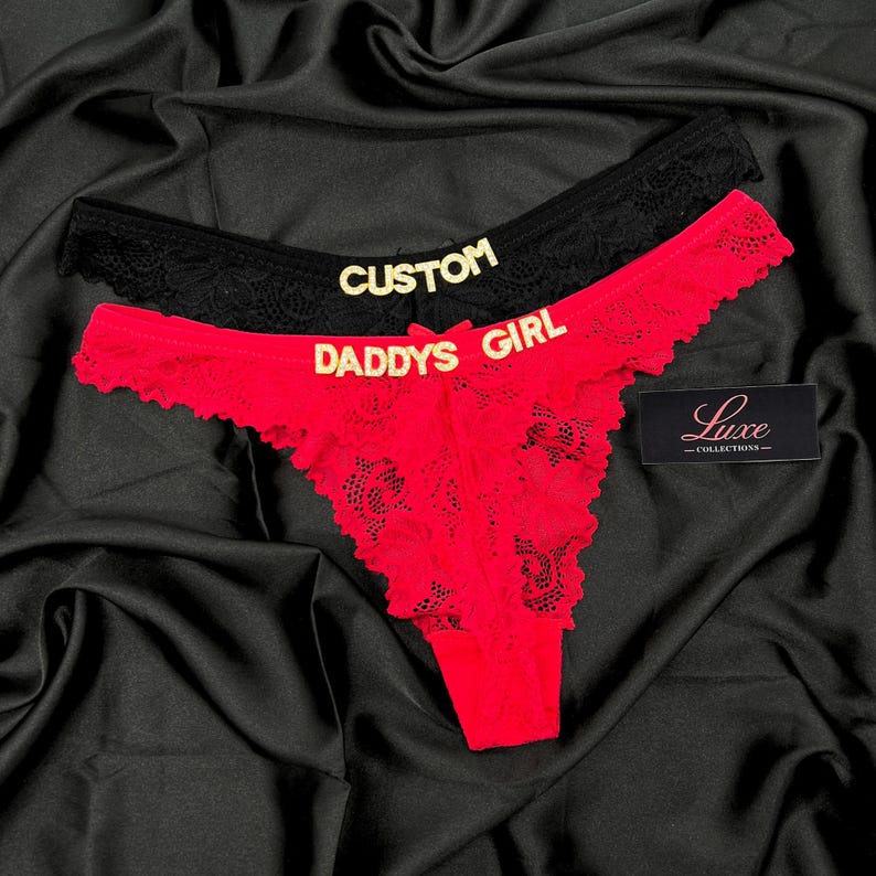Pode incluir: Um par de tangas de renda, uma preta e uma vermelha, com letras douradas. A pe&ccedil;a preta exibe a palavra CUSTOM, enquanto a pe&ccedil;a vermelha exibe o texto DADDYS GIRL. Uma pequena etiqueta preta com o texto branco Luxe COLLECTIONS &eacute; vis&iacute;vel.
