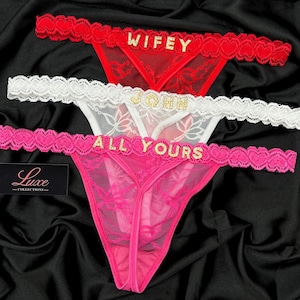 May include: Three pairs of lacy underwear with gold lettering. The top pair is red with the word "WIFEY" on it. The middle pair is white with the word "JOHN" on it. The bottom pair is pink with the words "ALL YOURS" on it.