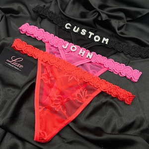 Puede incluir: Tres tangas en negro, rosa y rojo, cada uno con una cintura de encaje festoneado. El tanga negro muestra la palabra CUSTOM en pedrer&iacute;a plateada, mientras que el tanga rosa presenta el nombre JOHN en pedrer&iacute;a plateada en la cintura.