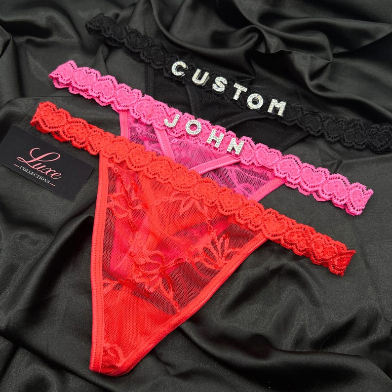 Puede incluir: Tres tangas en negro, rosa y rojo, cada uno con una cintura de encaje festoneado. El tanga negro muestra la palabra CUSTOM en pedrer&iacute;a plateada, mientras que el tanga rosa presenta el nombre JOHN en pedrer&iacute;a plateada en la cintura.