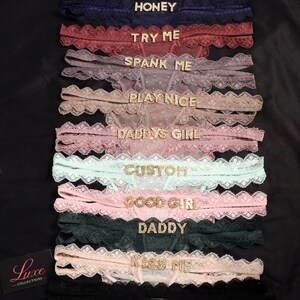 K&ouml;nnte beinhalten: Eine Kollektion von zehn Spitzen-Tangas in verschiedenen Farben, jeweils mit einem mit Strasssteinen verzierten Wort oder Satz am Bund. Der Text umfasst HONEY, TRY ME, SPANK ME, PLAY NICE, DADDYS GIRL, CUSTOM, GOOD GIRL, DADDY und KISS ME.