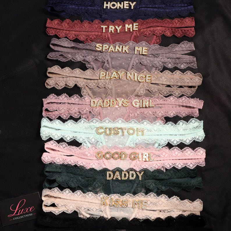 K&ouml;nnte beinhalten: Eine Kollektion von zehn Spitzen-Tangas in verschiedenen Farben, jeweils mit einem mit Strasssteinen verzierten Wort oder Satz am Bund. Der Text umfasst HONEY, TRY ME, SPANK ME, PLAY NICE, DADDYS GIRL, CUSTOM, GOOD GIRL, DADDY und KISS ME.