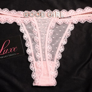 May include: A light pink thong featuring sheer dotted mesh fabric, scalloped lace trim, and a rhinestone embellishment on the waistband that reads GOOD GIRL. A black rectangular card with the white text Luxe COLLECTIONS is visible.