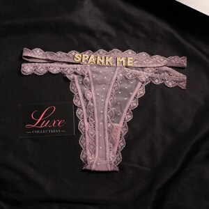 K&ouml;nnte beinhalten: Ein malvenfarbener Tanga aus transparentem Netzstoff mit Punkten, Spitzenbesatz und einem doppelten Bund, der mit goldfarbenen Strassbuchstaben verziert ist, die SPANK ME bilden. Eine schwarze Karte mit dem rosa Text Luxe COLLECTIONS liegt bei.