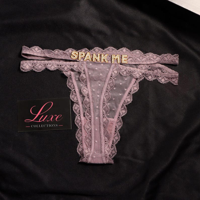K&ouml;nnte beinhalten: Ein malvenfarbener Tanga aus transparentem Netzstoff mit Punkten, Spitzenbesatz und einem doppelten Bund, der mit goldfarbenen Strassbuchstaben verziert ist, die SPANK ME bilden. Eine schwarze Karte mit dem rosa Text Luxe COLLECTIONS liegt bei.