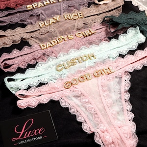 K&ouml;nnte beinhalten: Eine Kollektion aus Spitzen-Tangas in verschiedenen Pastellfarben wie Hellrosa, Mintgr&uuml;n und Mauve. Jedes St&uuml;ck ist mit einem dekorativen Strass-Schriftzug am Bund versehen, wie GOOD GIRL, CUSTOM, DADDYS GIRL, PLAY NICE und SPANK ME. Eine schwarze Karte mit dem Text Luxe COLLECTIONS ist zu sehen.
