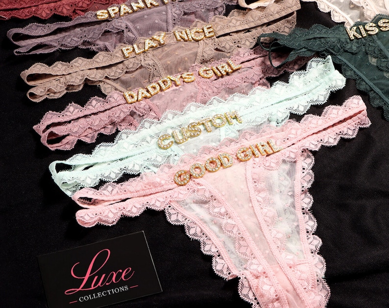 K&ouml;nnte beinhalten: Eine Kollektion aus Spitzen-Tangas in verschiedenen Pastellfarben wie Hellrosa, Mintgr&uuml;n und Mauve. Jedes St&uuml;ck ist mit einem dekorativen Strass-Schriftzug am Bund versehen, wie GOOD GIRL, CUSTOM, DADDYS GIRL, PLAY NICE und SPANK ME. Eine schwarze Karte mit dem Text Luxe COLLECTIONS ist zu sehen.