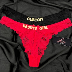 Op de afbeelding: Twee paar kanten ondergoed, een zwarte en een rode, met gouden letters. Het zwarte paar toont het woord CUSTOM, terwijl het rode paar de tekst DADDYS GIRL bevat. Een klein zwart rechthoekig label met de tekst Luxe COLLECTIONS is inbegrepen.