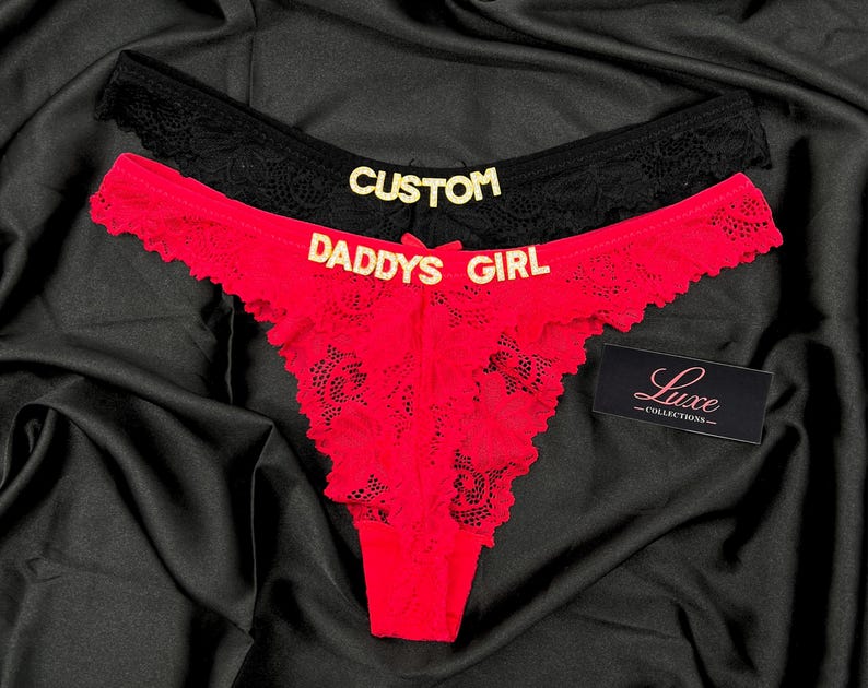 Pode incluir: Dois pares de roupa interior de renda, um preto e um vermelho, com letras douradas. O par preto exibe a palavra CUSTOM, enquanto o par vermelho apresenta o texto DADDYS GIRL. Inclui uma pequena etiqueta preta com o texto Luxe COLLECTIONS.