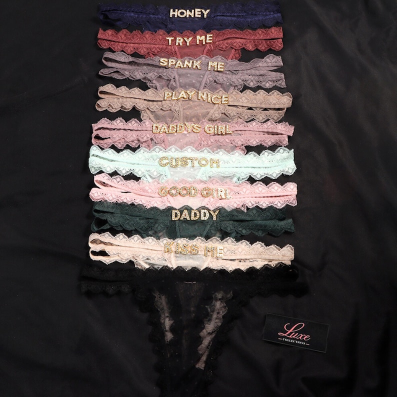 K&ouml;nnte beinhalten: Eine Kollektion von zehn Spitzen-Tangas in verschiedenen Farben wie Marineblau, Rot, Mauve, Beige, Rosa, Mint und Schwarz. Jeder Tanga ist mit goldfarbenen Buchstaben verziert, die W&ouml;rter wie HONEY, TRY ME, SPANK ME, PLAY NICE, DADDY'S GIRL, CUSTOM, GOOD GIRL, DADDY und KISS ME bilden.